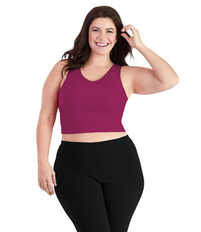 Plus size woman, facing front, wearing JunoActive plus size Stretch Naturals V-Neck Bra top in Merlot. The woman is wearing black JunoActive plus size leggings. Her left arm is tucking her hair behind her ear, her right arm falls naturally to her side.