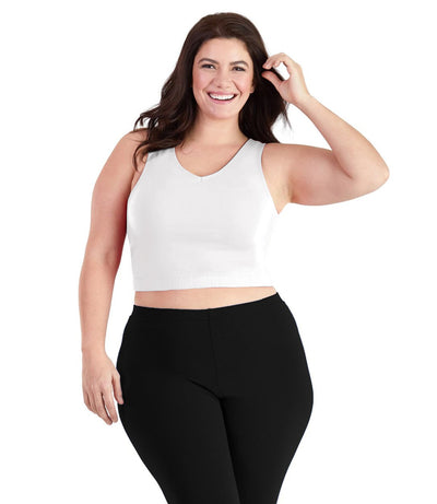 Plus size woman, facing front, wearing JunoActive plus size Stretch Naturals V-Neck Bra top in white. The woman is wearing black JunoActive plus size leggings. Her left arm is up tucking her hair behind her ear, her right arm falls naturally to her side.