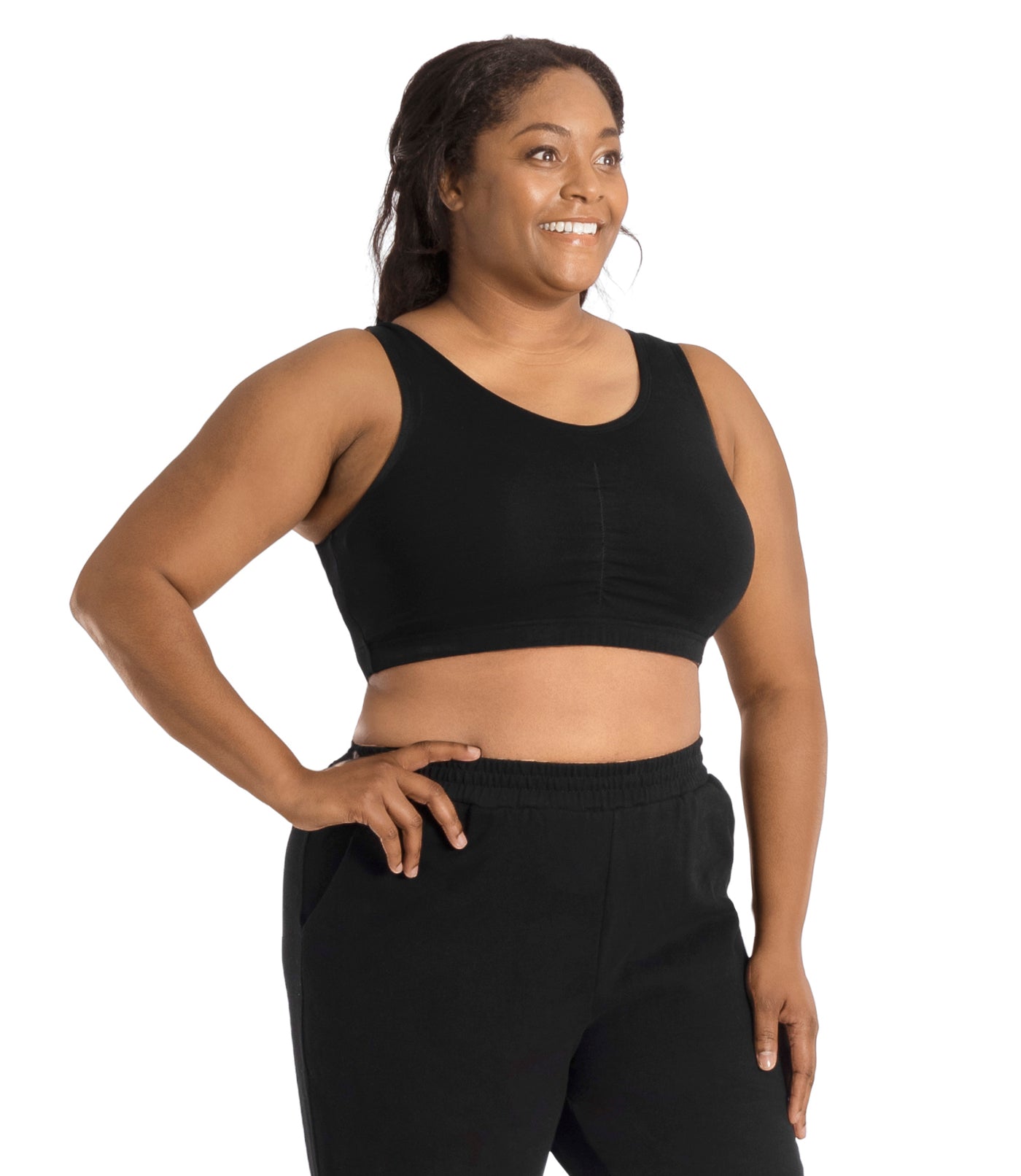 Plus size woman, facing to the side, looking away, right hand on hip, left hand by side, wearing JunoActive Stretch Naturals Shirred Bra Top. Bra is gathered in middle and is a scoop top neck. Straps are wide and bra color is black.