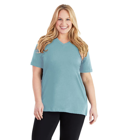 Plus size woman, facing front, wearing JunoActive plus size Stretch Naturals V-Neck in the color Soft Green. She is wearing JunoActive Plus Size Leggings in the color black. 