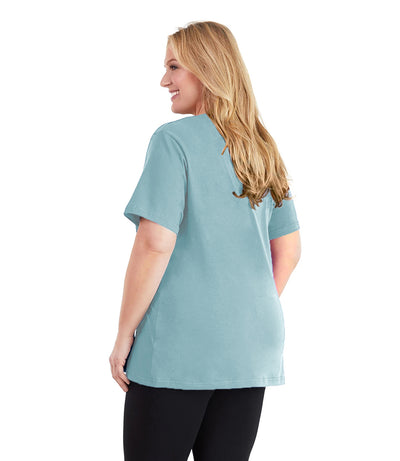Plus size woman, facing back and looking left, wearing JunoActive plus size Stretch Naturals V-Neck in the color Soft Green. She is wearing JunoActive Plus Size Leggings in the color black.