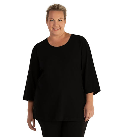Plus size woman, facing front, wearing JunoActive plus size Stretch Naturals Scoop Neck ¾ Sleeve Top in the color Black. She is wearing JunoActive Plus Size Leggings in the color Black.