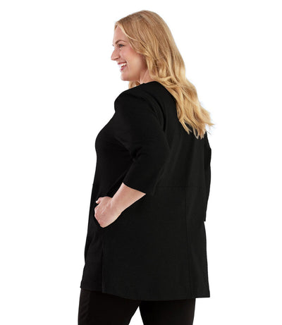 Plus size woman, facing back looking left, wearing JunoActive plus size Stretch Naturals Empire Tunic with Pockets in the color Black. Her left hand is in the tunic pocket at her waist level. She is wearing JunoActive Plus Size Leggings in the color Black.