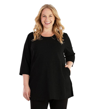 Plus size woman, facing front, wearing JunoActive plus size Stretch Naturals Empire Tunic with Pockets in the color Black. Her left hand is in the tunic pocket at her waist level. Her right hand hangs naturally at her side. She is wearing JunoActive Plus Size Leggings in the color Black.