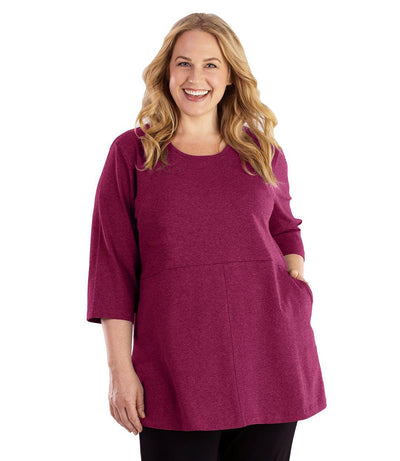 Plus size woman, facing front, wearing JunoActive plus size Stretch Naturals Empire Tunic with Pockets in the color Merlot. Her left hand is in the tunic pocket at her waist level. Her right hand hangs naturally at her side. She is wearing JunoActive Plus Size Leggings in the color Black.