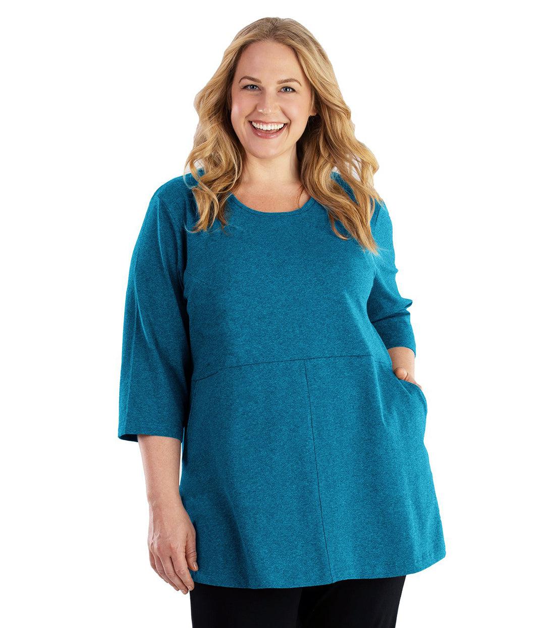 Plus size woman, facing front, wearing JunoActive plus size Stretch Naturals Empire Tunic with Pockets in the color Deep Teal. Her left hand is in the tunic pocket at her waist level. Her right hand hangs naturally at her side. She is wearing JunoActive Plus Size Leggings in the color Black.