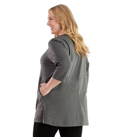 Plus size woman, facing back looking left, wearing JunoActive plus size Stretch Naturals Empire Tunic with Pockets in the color Heather Charcoal. Her left hand is in the tunic pocket at her waist level. She is wearing JunoActive Plus Size Leggings in the color Black.