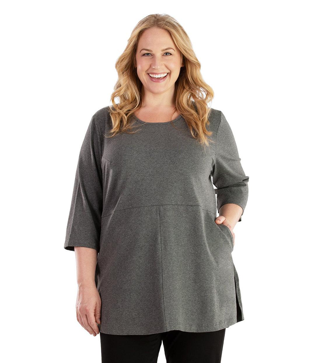 Plus size woman, facing front, wearing JunoActive plus size Stretch Naturals Empire Tunic with Pockets in the color Heather Charcoal. Her left hand is in the tunic pocket at her waist level. Her right hand hangs naturally at her side. She is wearing JunoActive Plus Size Leggings in the color Black.