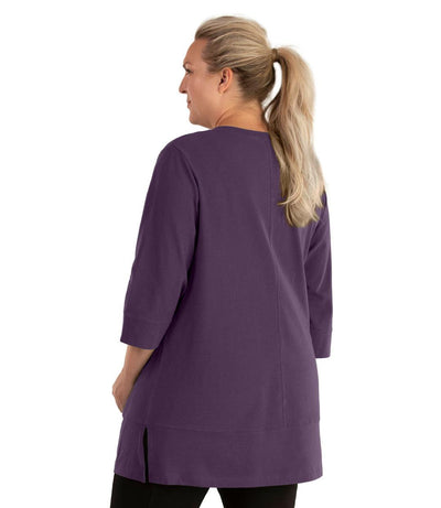 Plus size woman, facing back looking left, wearing JunoActive plus size Stretch Naturals Princess V-neck Tunic in the color Blackberry. She is wearing JunoActive Plus Size Leggings in the color Black.