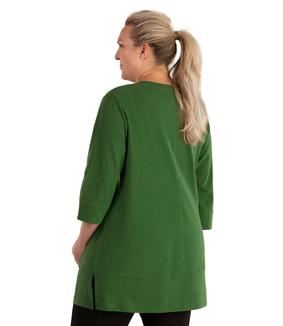 Plus size woman, facing back looking left, wearing JunoActive plus size Stretch Naturals Princess V-neck Tunic in the color Evergreen. She is wearing JunoActive Plus Size Leggings in the color Black.