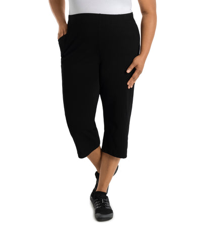 Plus size woman is facing forward, right hand in pocket, left arm and hand to side, wearing JunoActive Stretch Naturals side pocket plus size capri pant bottom. The hemline comes to mid-calf and hugs the body without being skin tight. Color is black.