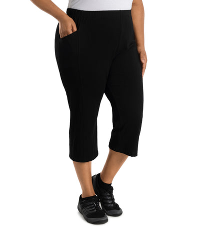 Plus size woman is facing at a side angle, right hand in pocket, left arm and hand to side, wearing JunoActive Stretch Naturals side pocket plus size capri pant bottom. The hemline comes to mid-calf and hugs the body without being skin tight. Color is black.