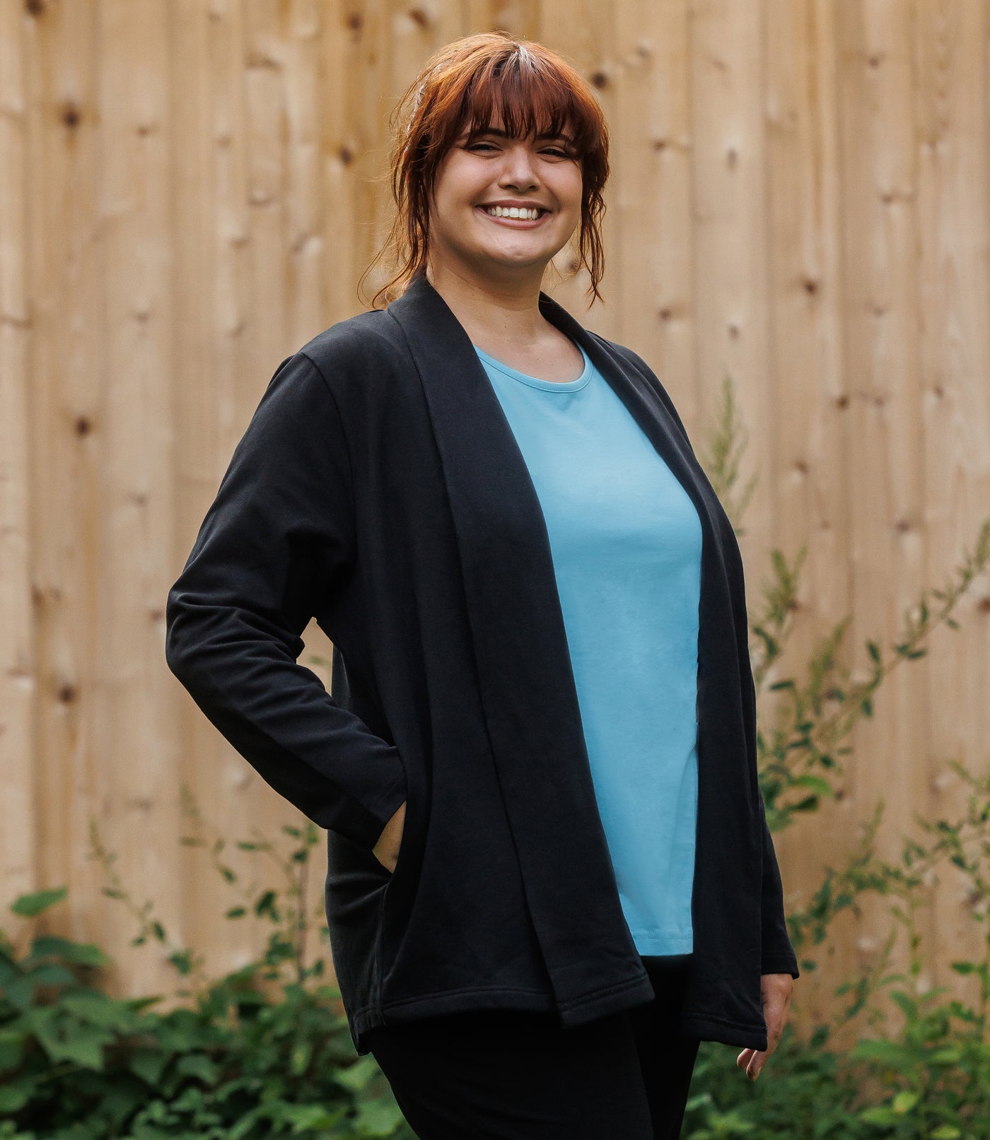Plus-size model, facing forward, wearing JunoActive's Mavie Cotton Wrap Jacket in color black. Her right hand is in her pocket of jacket and left hand hanging by her side. 