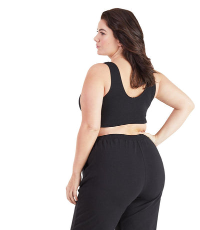 Plus size woman, facing back, wearing JunoActive plus size Stretch Naturals Scoop Neck Bra in black. The woman is wearing black plus size JunoActive loose fit leggings. Her left arm falls naturally to her side, her right hand is on her hip. 