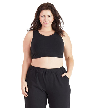Plus size woman, facing front, wearing JunoActive plus size Stretch Naturals Scoop Neck Bra in black. The woman is wearing black plus size JunoActive loose fit leggings. Her right arm falls naturally to her side, her left arm is in the legging pocket. 
