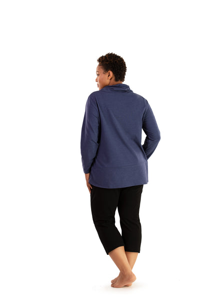 Plus size woman, facing back looking left, wearing JunoActive plus size Stretch Naturals Cowl Top in the color Denim Blue. She is wearing JunoActive Plus Size Capri Leggings in the color Black.