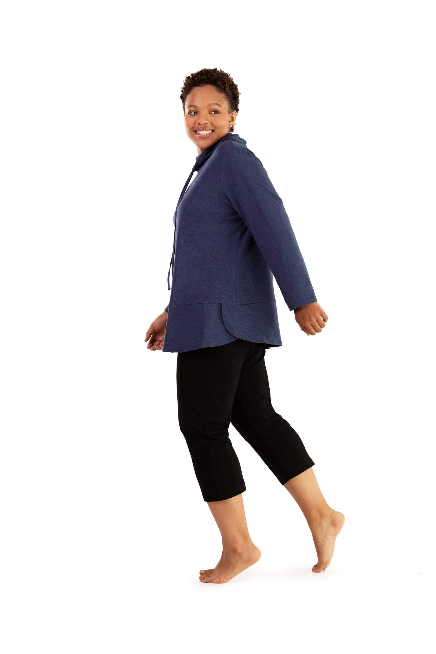 Plus size woman, walking back left looking forward, wearing JunoActive plus size Stretch Naturals Cowl Top in the color Denim Blue. She is wearing JunoActive Plus Size Capri Leggings in the color Black.