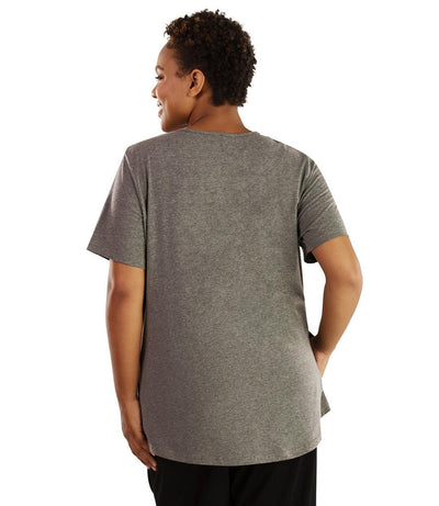 Plus size woman, facing back looking left, wearing JunoActive plus size Stretch Naturals Lite Pocket Tee in the color Heather Grey. Her right hand is in the shirt pocket by her hip, her left arm is naturally at her side. She is wearing JunoActive Plus Size Leggings in the color Black.