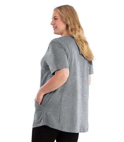 Plus size woman, facing back looking left, wearing JunoActive plus size Stretch Naturals Lite Pocketed V-Neck Short Sleeve Top in the color Heather Grey. Her left hand is in the shirt pocket by her hip. She is wearing JunoActive Plus Size Leggings in the color Black.