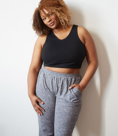 Plus size woman, front view, wearing a black JunoActive plus size sports bra in the color black and JunoActive Softwik Pocketed Pant in color heather grey. Her left hand is in the pant pocket at her hip, her right arm falls naturally at her side.